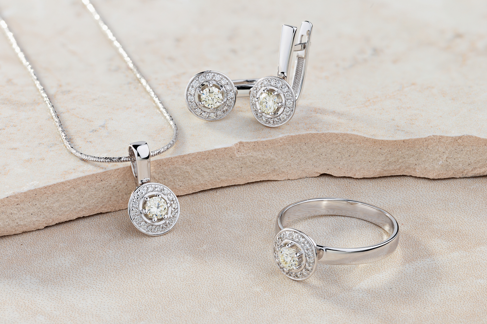 White gold necklace, earrings, and engagement ring all set with diamonds on stone surface.