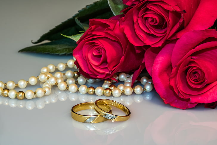 Two wedding bands stacked in front of pearls and red roses.
