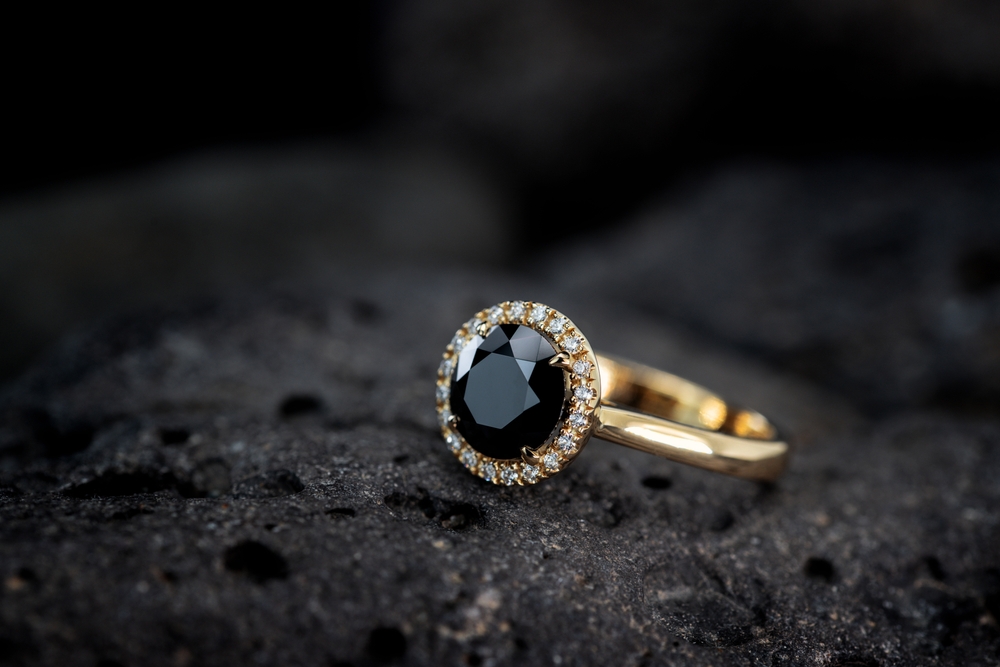 Yellow gold ring centered with a black diamond surrounded by a halo of white diamonds on a black fabric background.