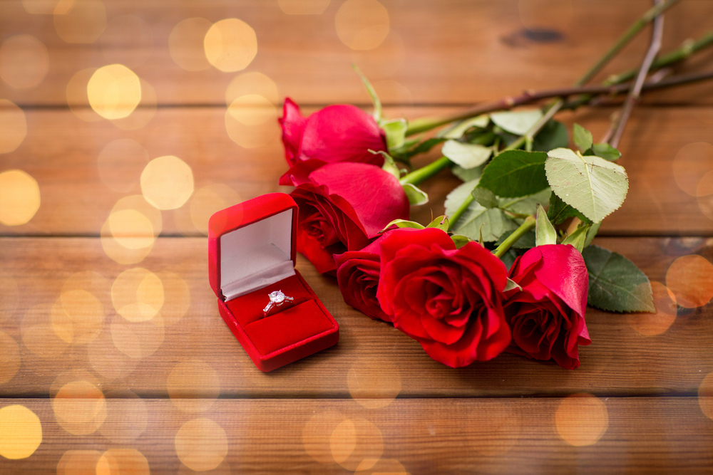 Engagement ring in red ring box next to red roses on a wooden table.
