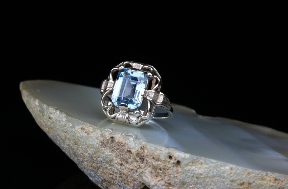 White gold filigree engagement ring set with a light blue gemstone on a slab on granite.