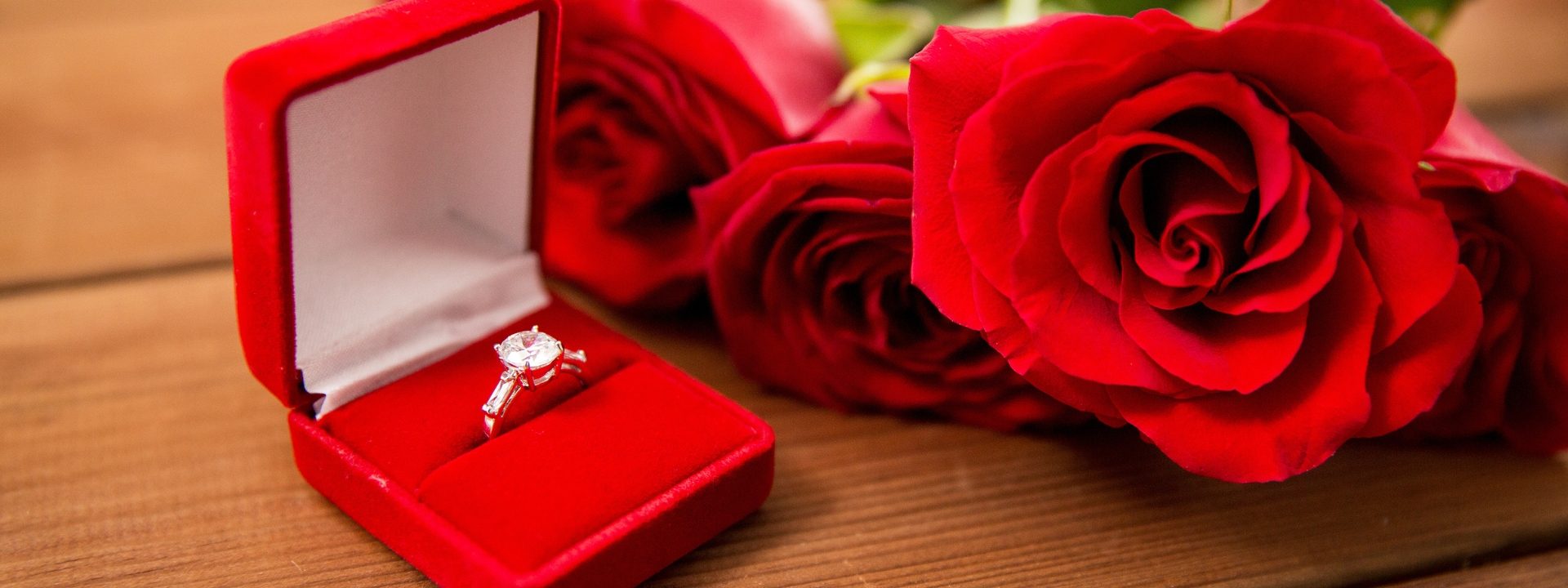 White gold three-stone diamond engagement ring in a red ring box next to red roses.