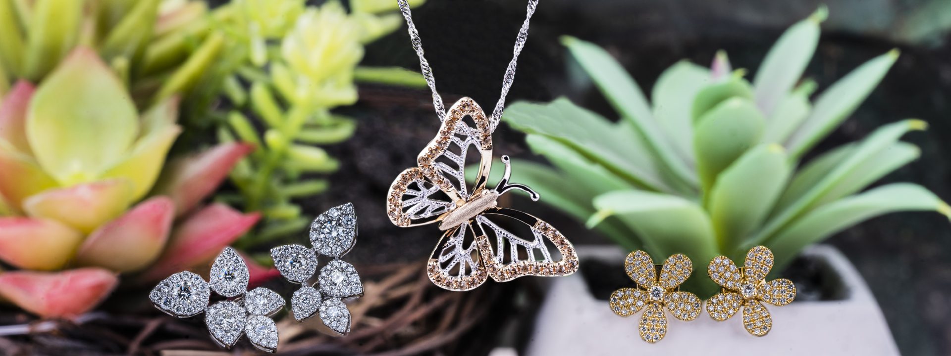 White gold diamond leaf earrings, yellow gold flower stud earrings, and rose and white gold diamond butterfly pendant in front of succulents.
