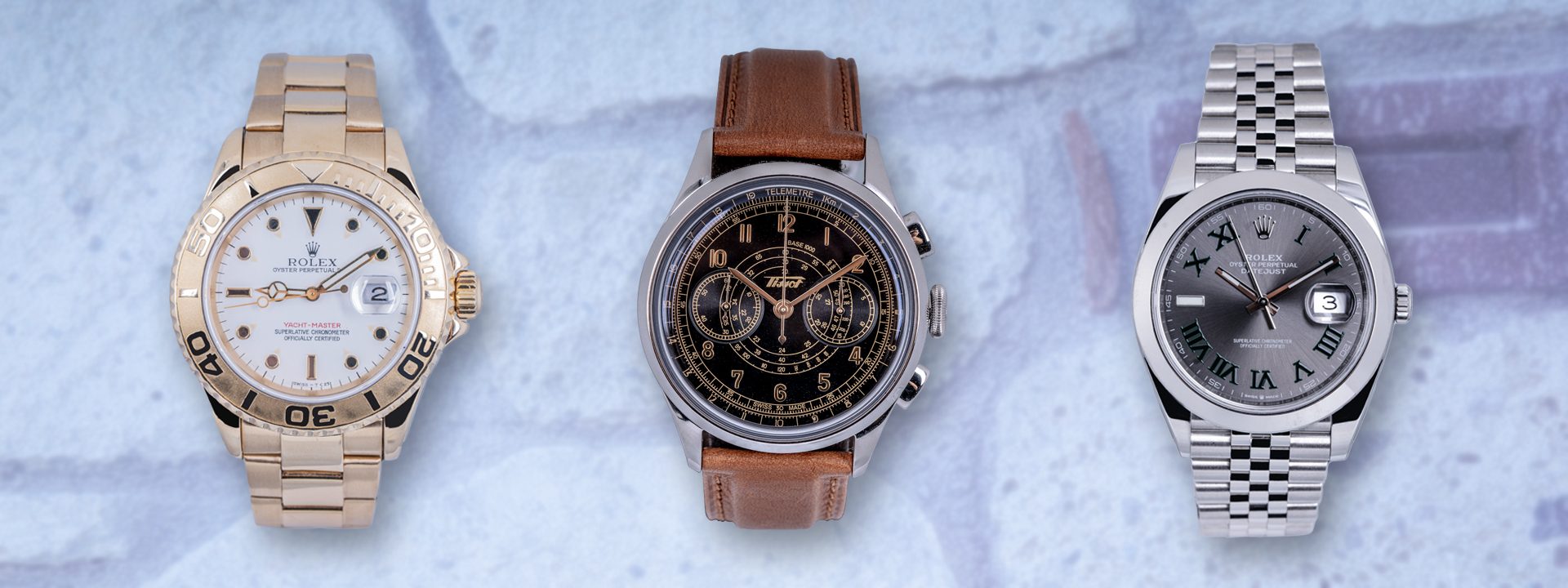 Three pre-owned watches including two Rolex watches and one Tissot in front of brick wall background.