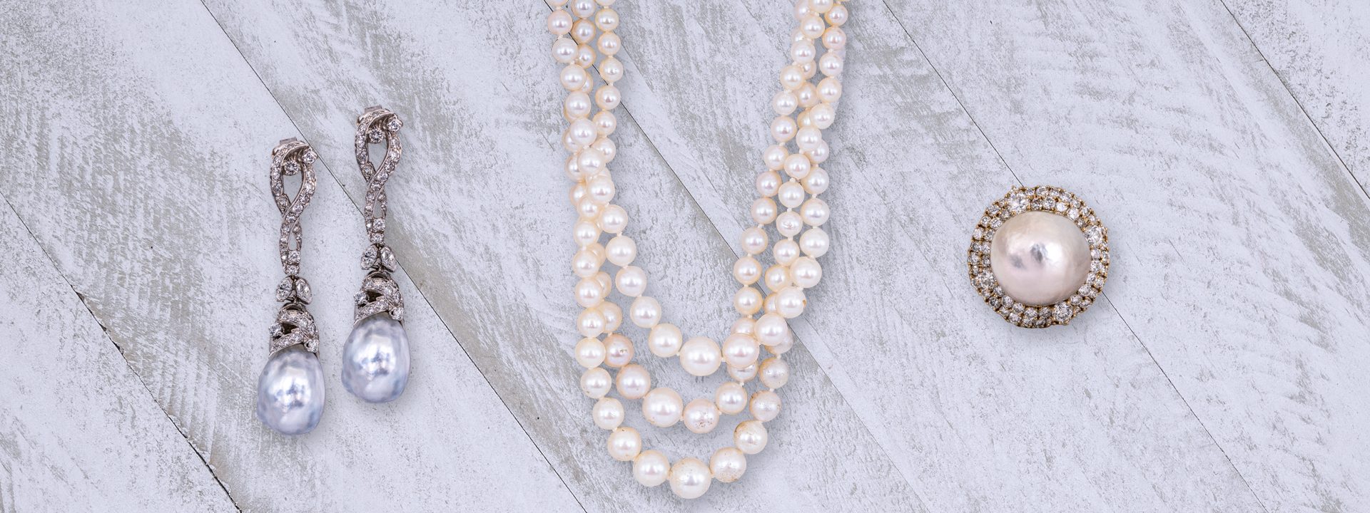 Pearl drop earrings, pearl cocktail ring, and three-strand pearl necklace on a wooden plank background.