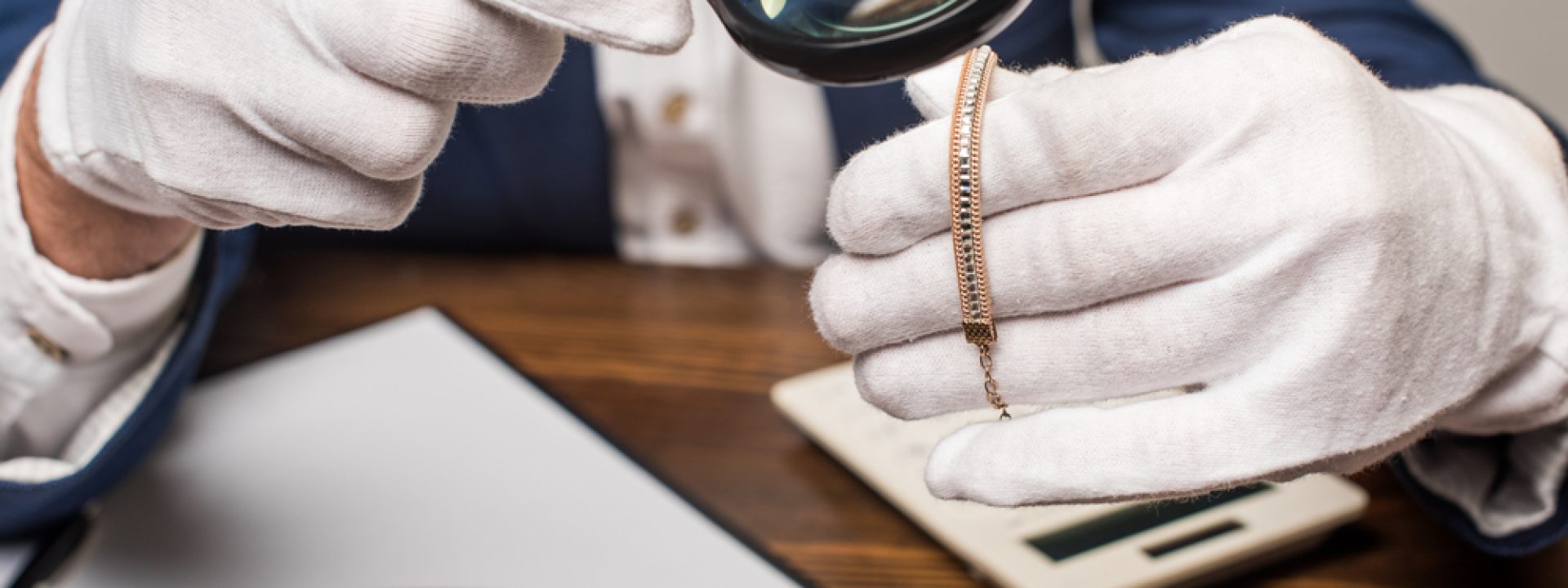 an appraiser wearing white gloves using a magnifying glass to inspect a gold and diamond bracelet